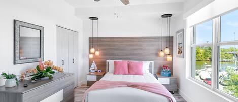 Un dormitorio moderno con cama, ventilador de techo, lámparas colgantes, cómoda, mesita de noche y ventana con vista a barcos.