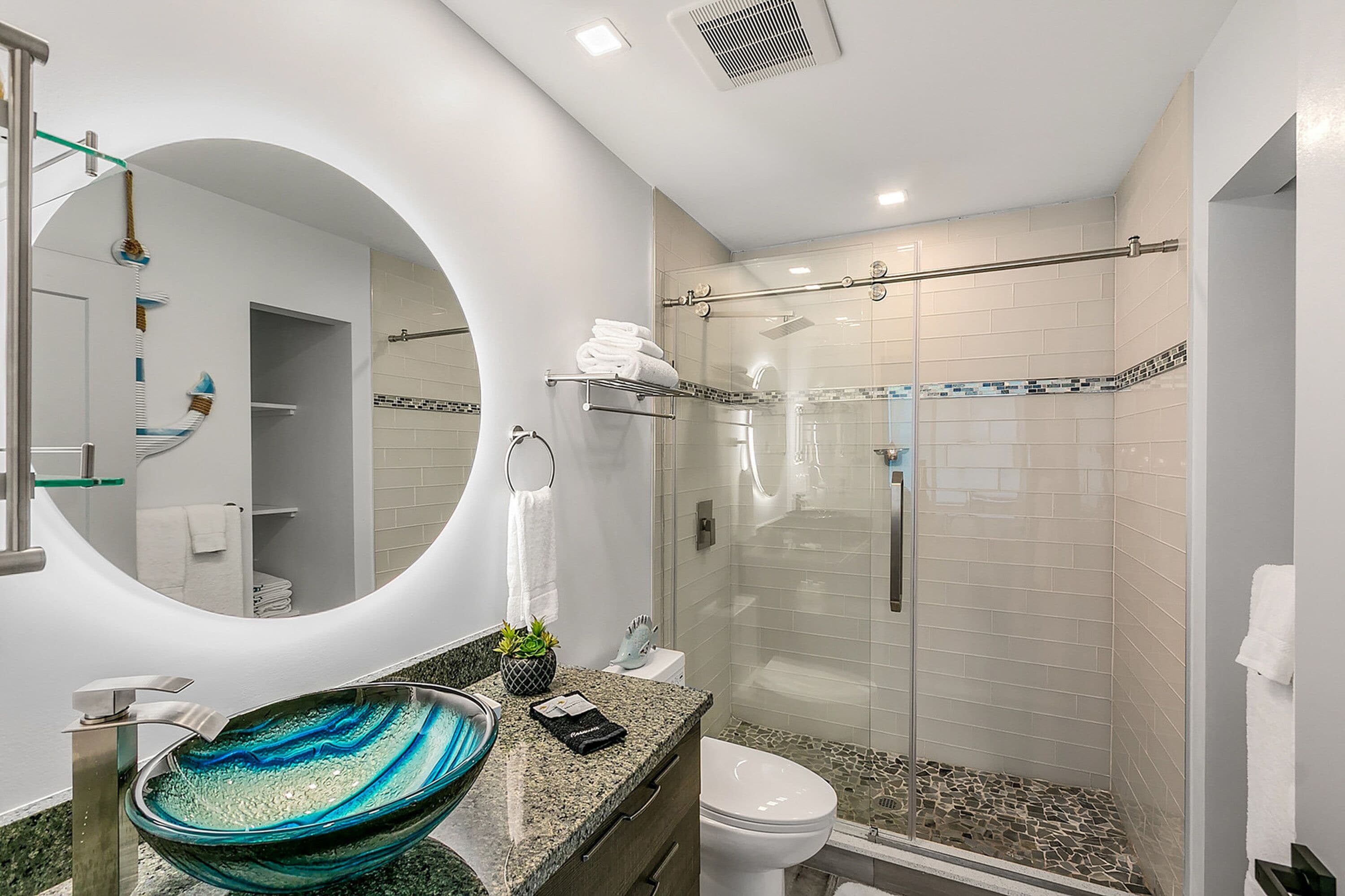 A modern bathroom with a glass-enclosed shower, a round mirror, a glass bowl sink, and a toilet.