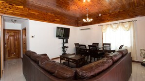 A living room with a brown leather sofa, a dining table with chairs, a TV mounted on the wall, and a wooden ceiling. - New Kingston Guest Apt at Fairway (Kingston)