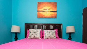 A room with a bright blue wall, a pink bedspread, and a sunset painting above the bed. - The Hellshire Beach House (Hellshire)