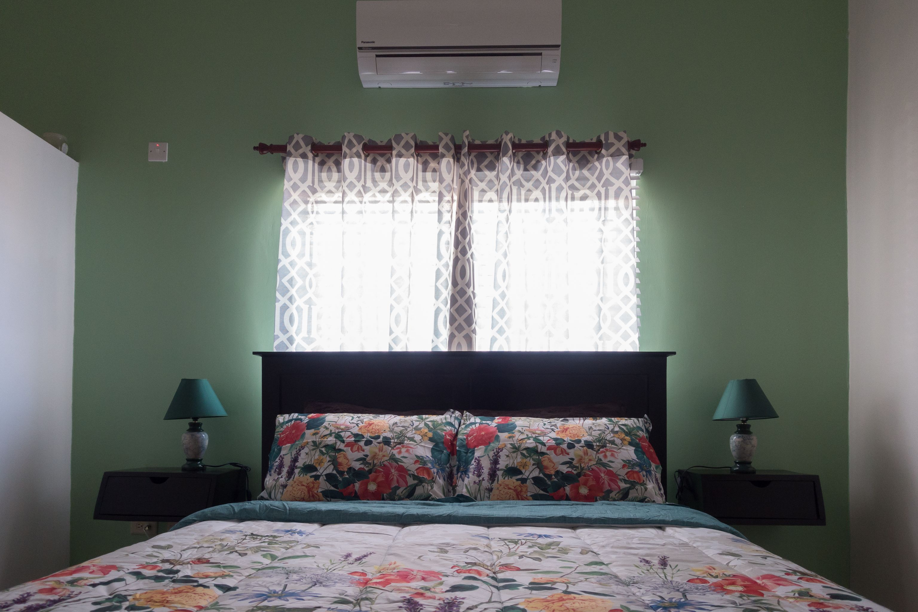A bedroom with a bed, two nightstands, a window with patterned curtains, and an air conditioner on the wall.