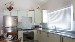 A kitchen with white cabinets, stainless steel appliances, a granite countertop, and a dining area with a wooden table and chairs. - The Country Club Villa I (Kingston)