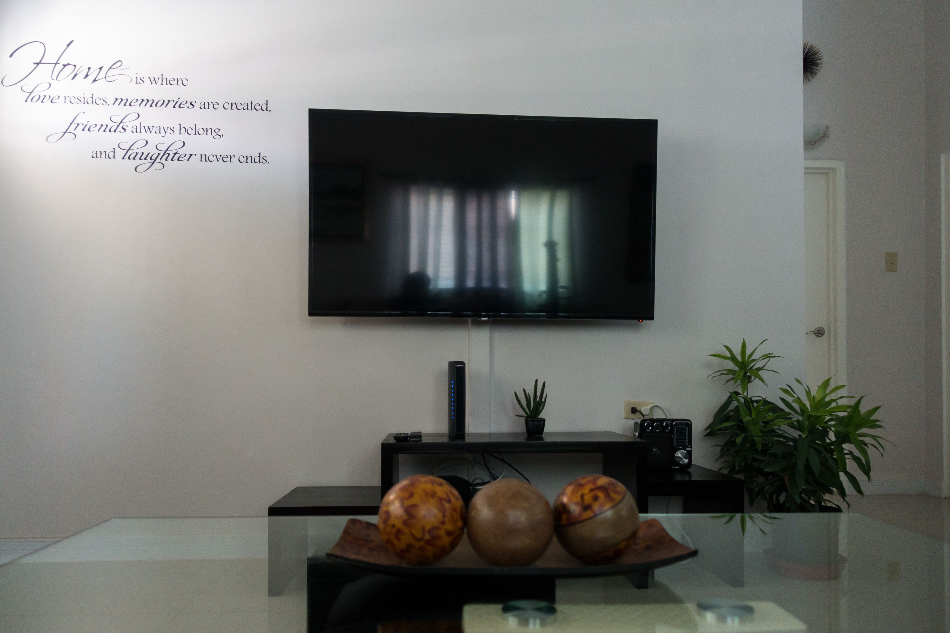 A living room with a flat-screen TV mounted on the wall, a glass coffee table with decorative bowls, a black entertainment unit below the TV, and a potted plant to the right.