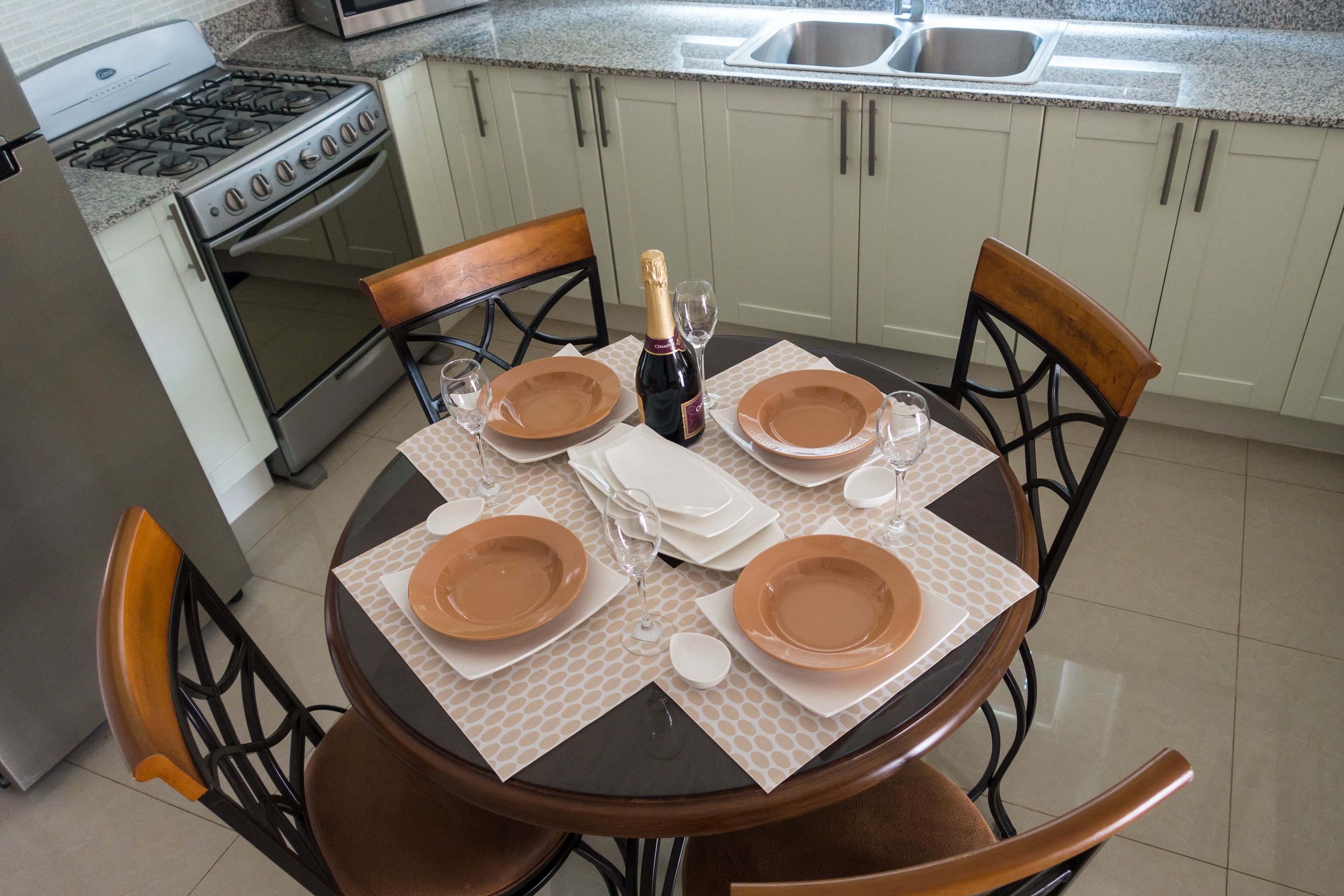 A dining table set with plates, glasses, and a bottle of champagne in a kitchen with white cabinets and a stainless steel sink.