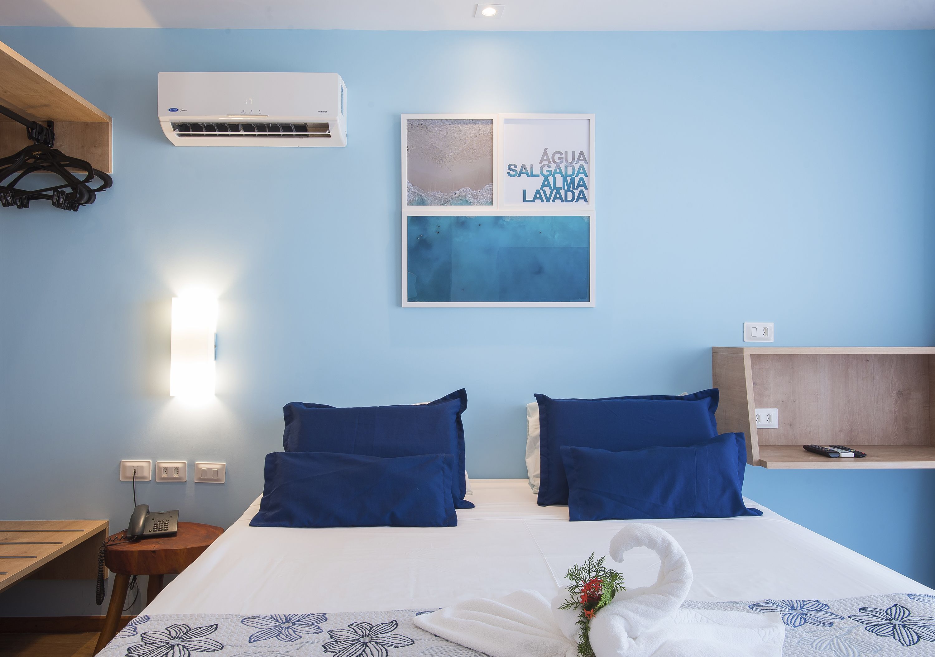 A hotel room with a bed, two blue pillows, a wall-mounted air conditioner, a framed artwork, a bedside table with a phone, and a shelf with a remote control.