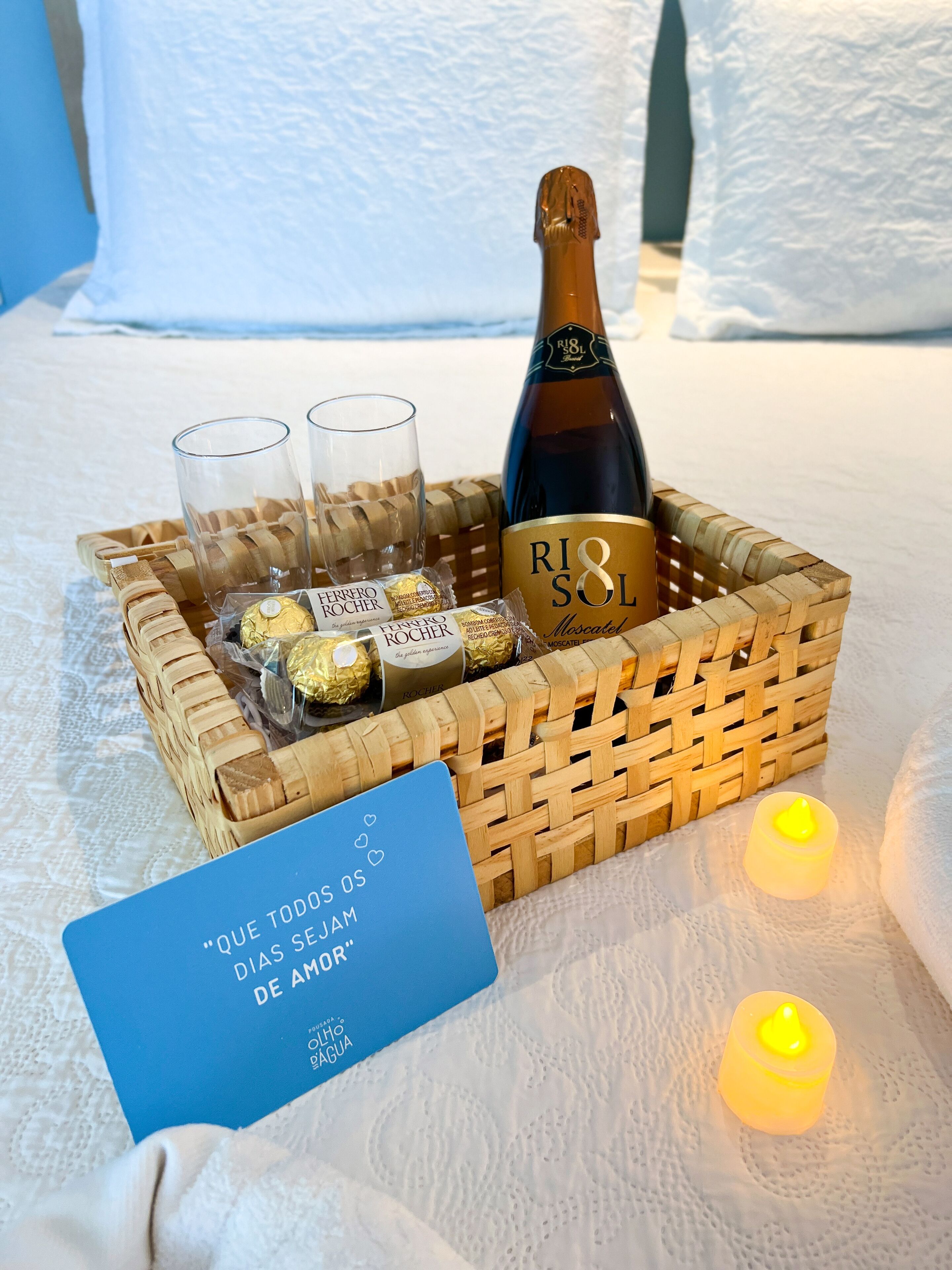 Una cesta de mimbre con una botella de champán Riosol Mascatel, dos copas y bombones Ferrero Rocher sobre una cama con almohada blanca y una vela encendida.