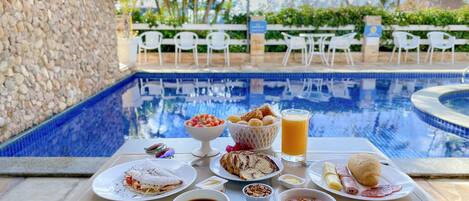 Une table de petit-déjeuner avec divers aliments et une tasse de café, placée au bord d’une piscine.