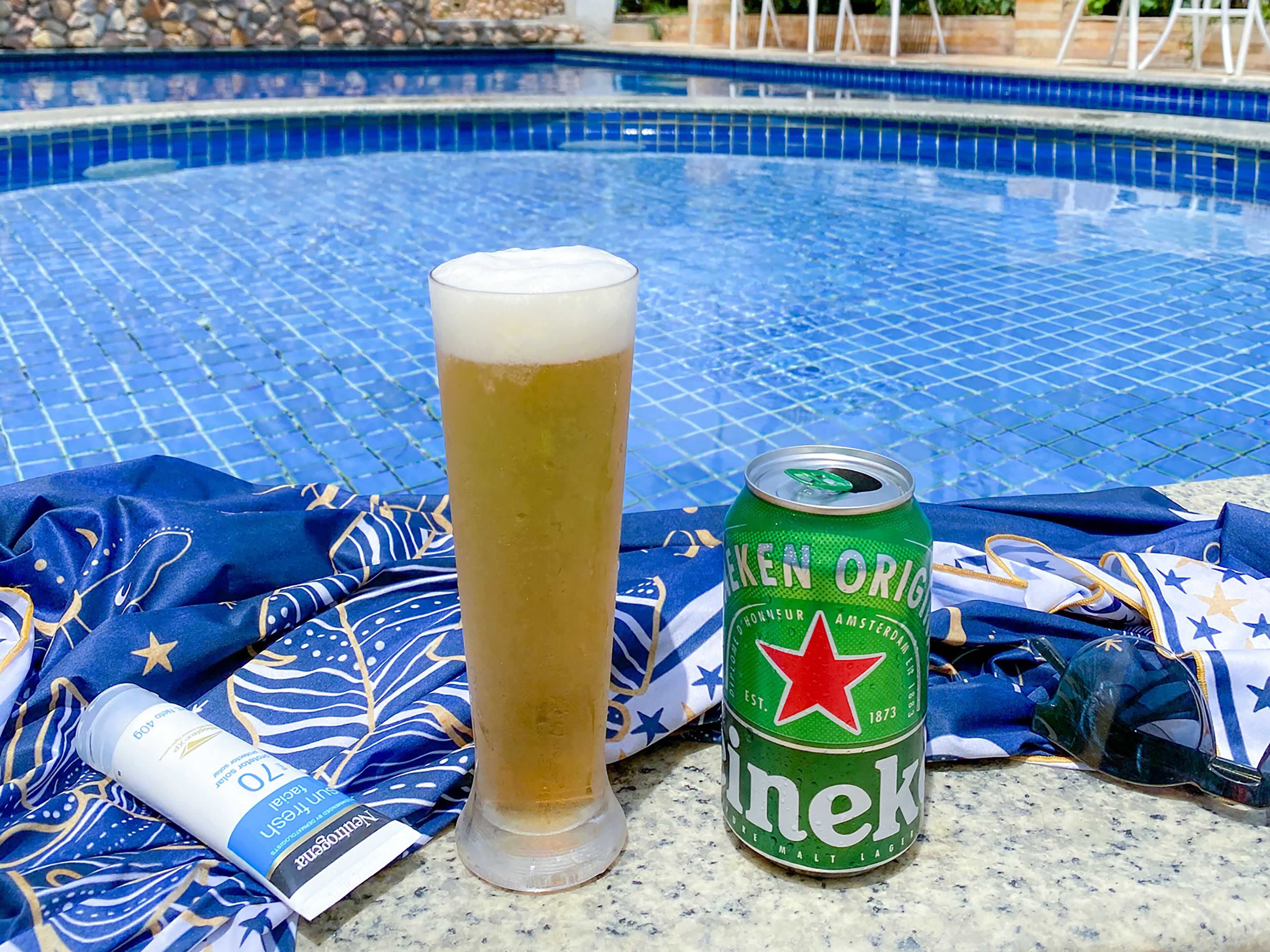 A glass of beer and a can of Heineken beer by a poolside.