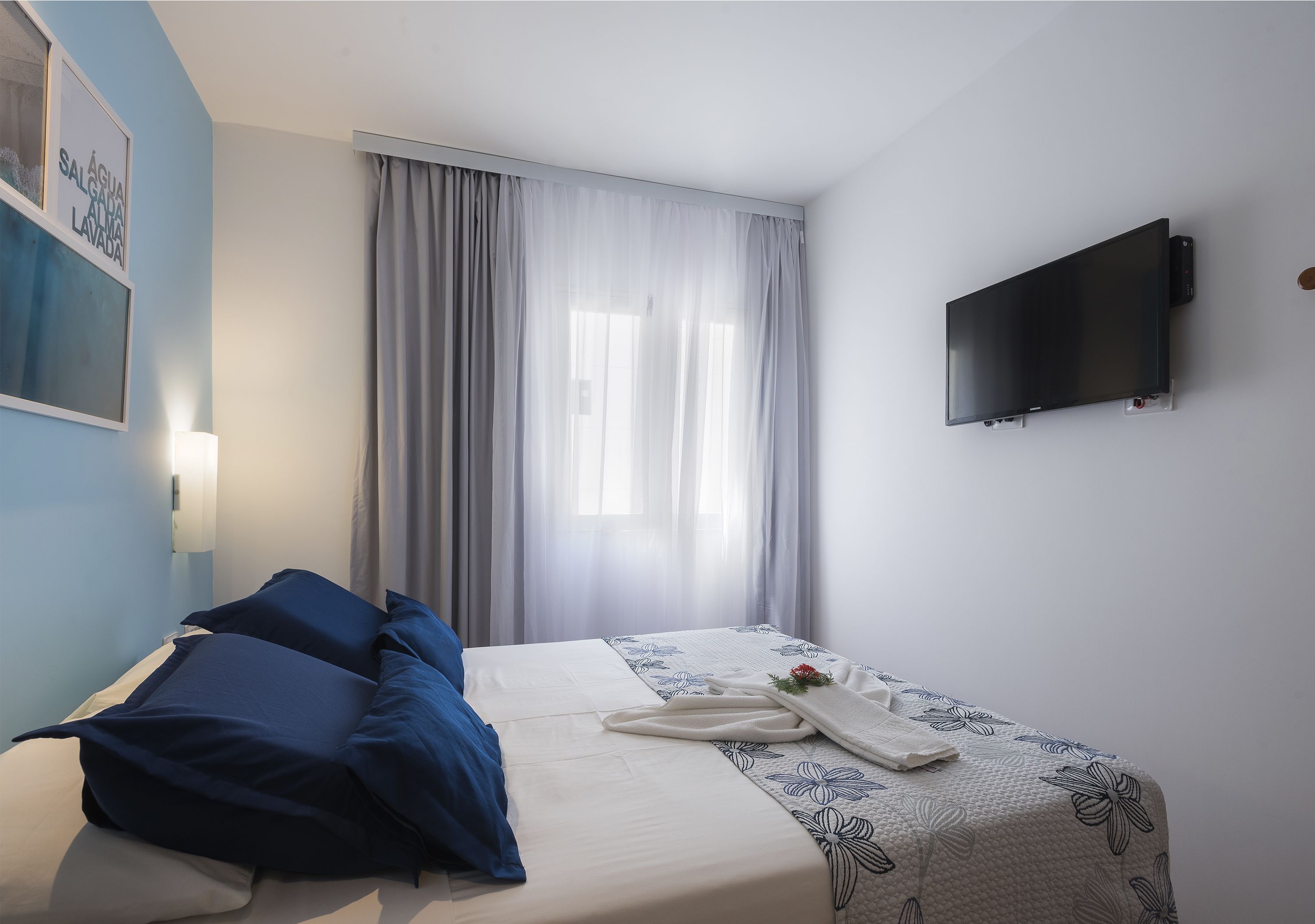 A hotel room with a bed, a flat-screen TV mounted on the wall, and a window with curtains.
