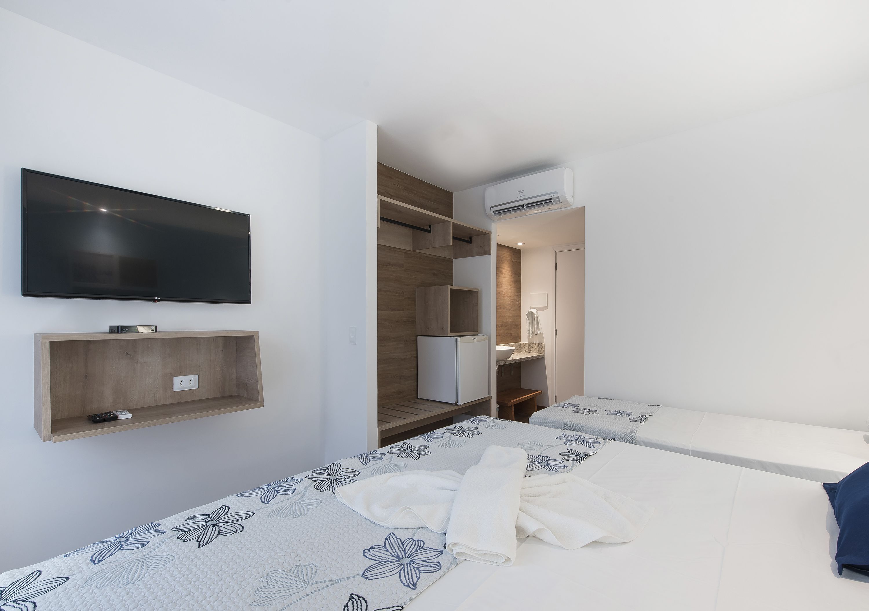 Una habitación de hotel moderna con una televisión de pantalla plana, un estante de madera, un mini refrigerador, un escritorio y una cama con ropa de cama estampada con motivos florales.
