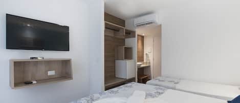 A modern hotel room with a flat-screen TV, a wooden shelf, a mini-fridge, a desk, and a bed with floral bedding.