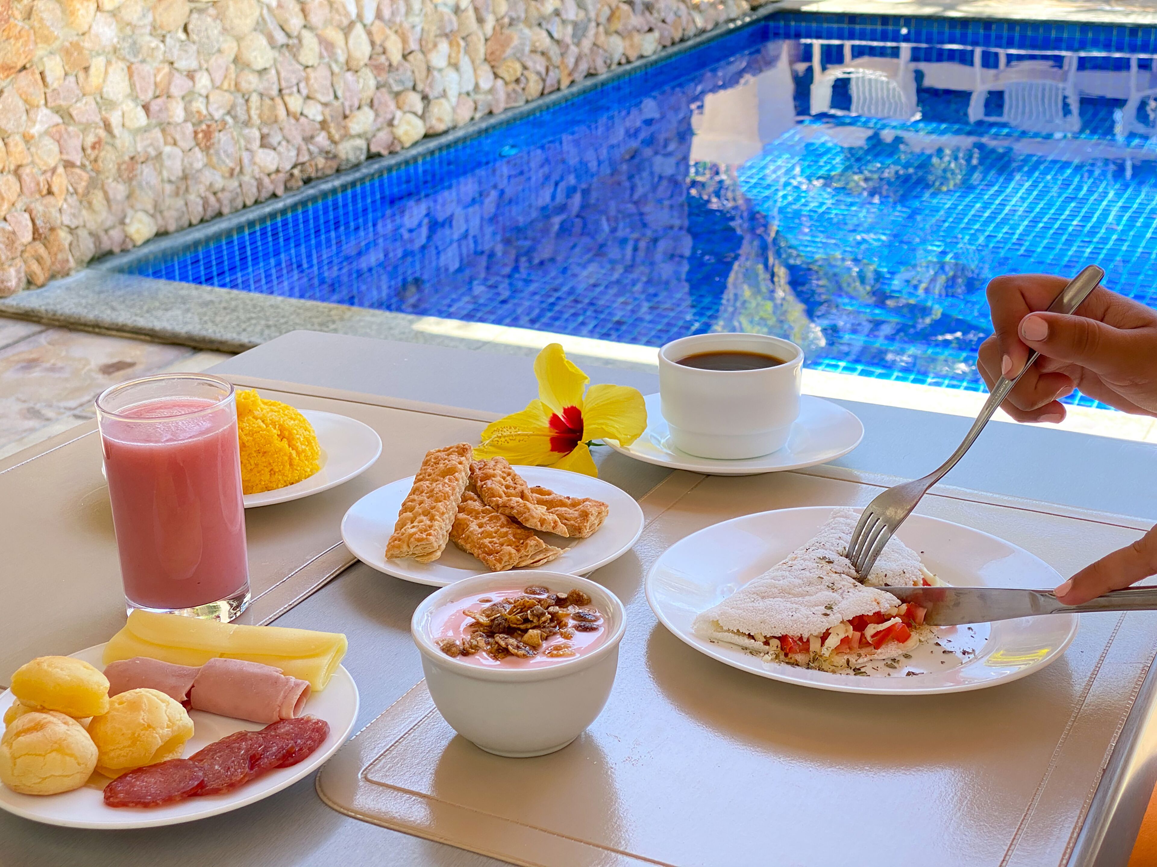 A breakfast table with various food items and a cup of coffee, overlooking a pool.