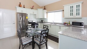 A modern kitchen with white cabinets, a stainless steel refrigerator, a microwave, and a dining area with a glass table and chairs. - The Country Club Villa II (Kingston)