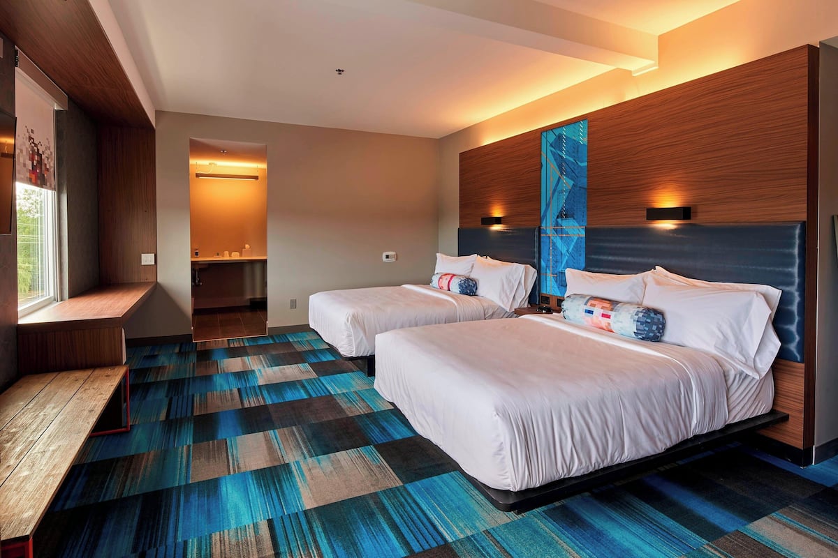 A modern hotel room with two beds, a bench, and a view of the outdoors.