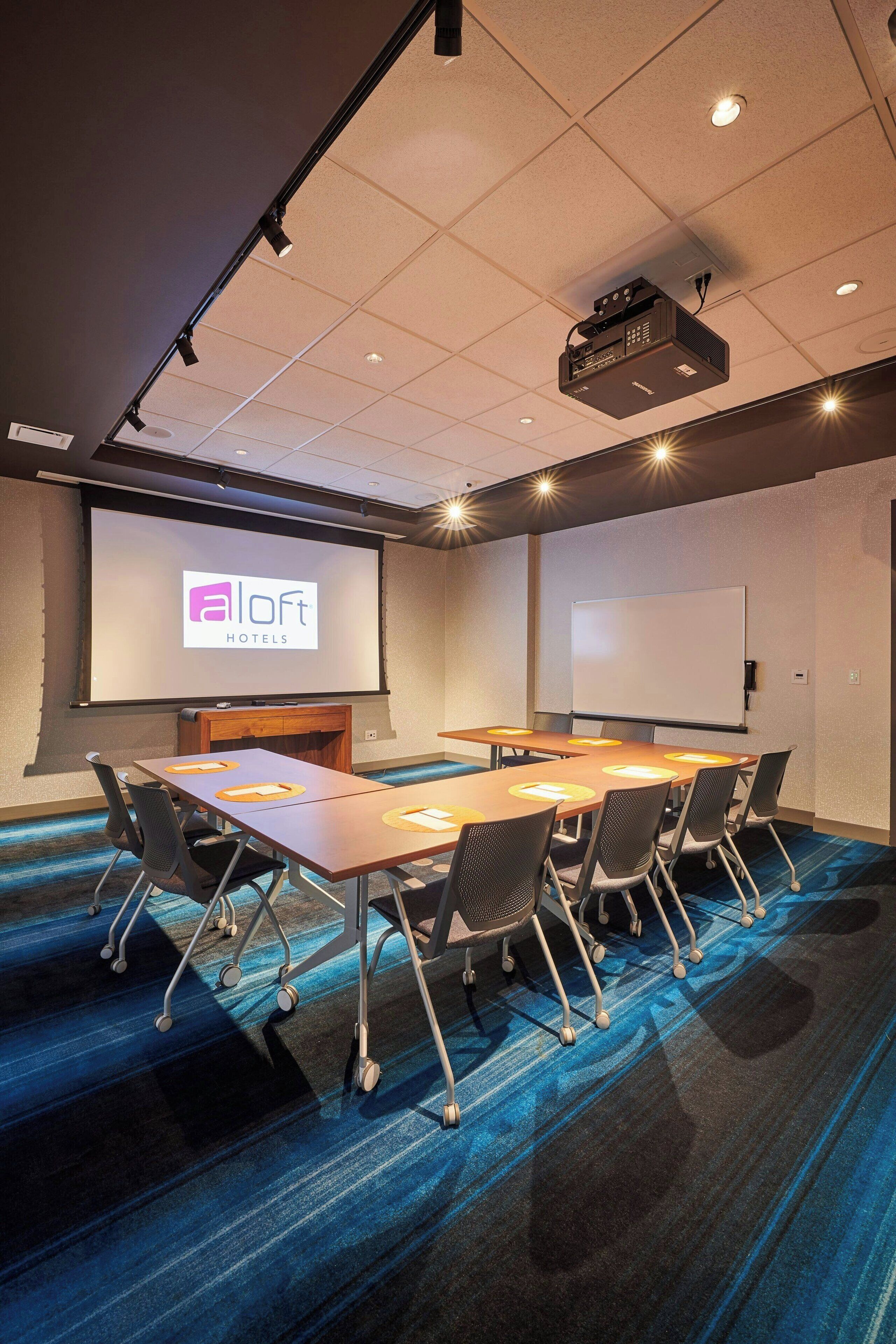 A conference room with a large screen displaying 'Aloft Hotels' logo, a whiteboard, and a projector. The room has a carpeted floor and a long table with chairs.