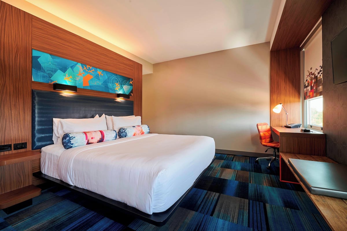 A modern hotel room with a large bed, a desk with a laptop, a chair, and a painting on the wall.