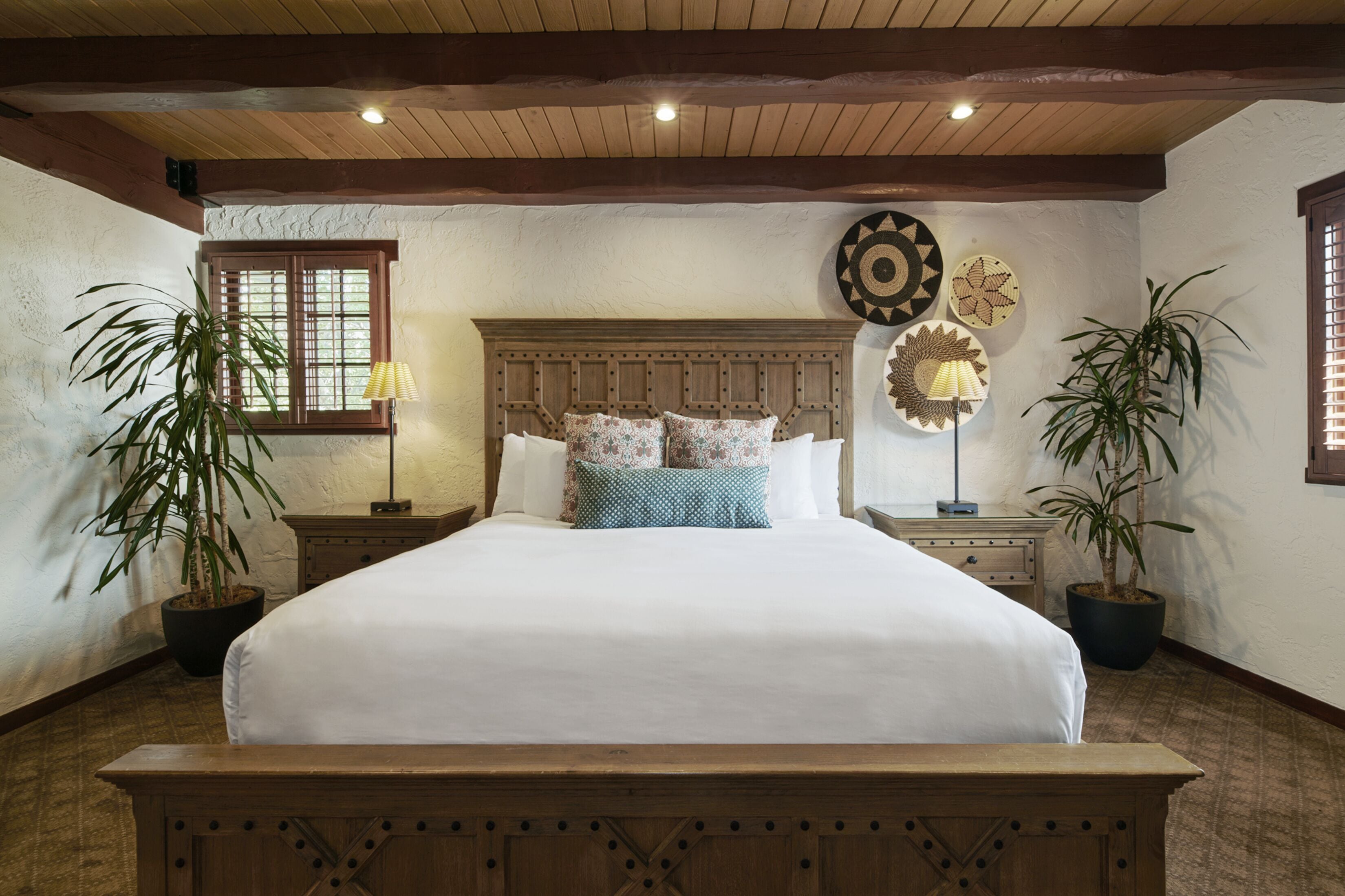 A bedroom with a wooden bed, two nightstands, a window with blinds, and two potted plants.