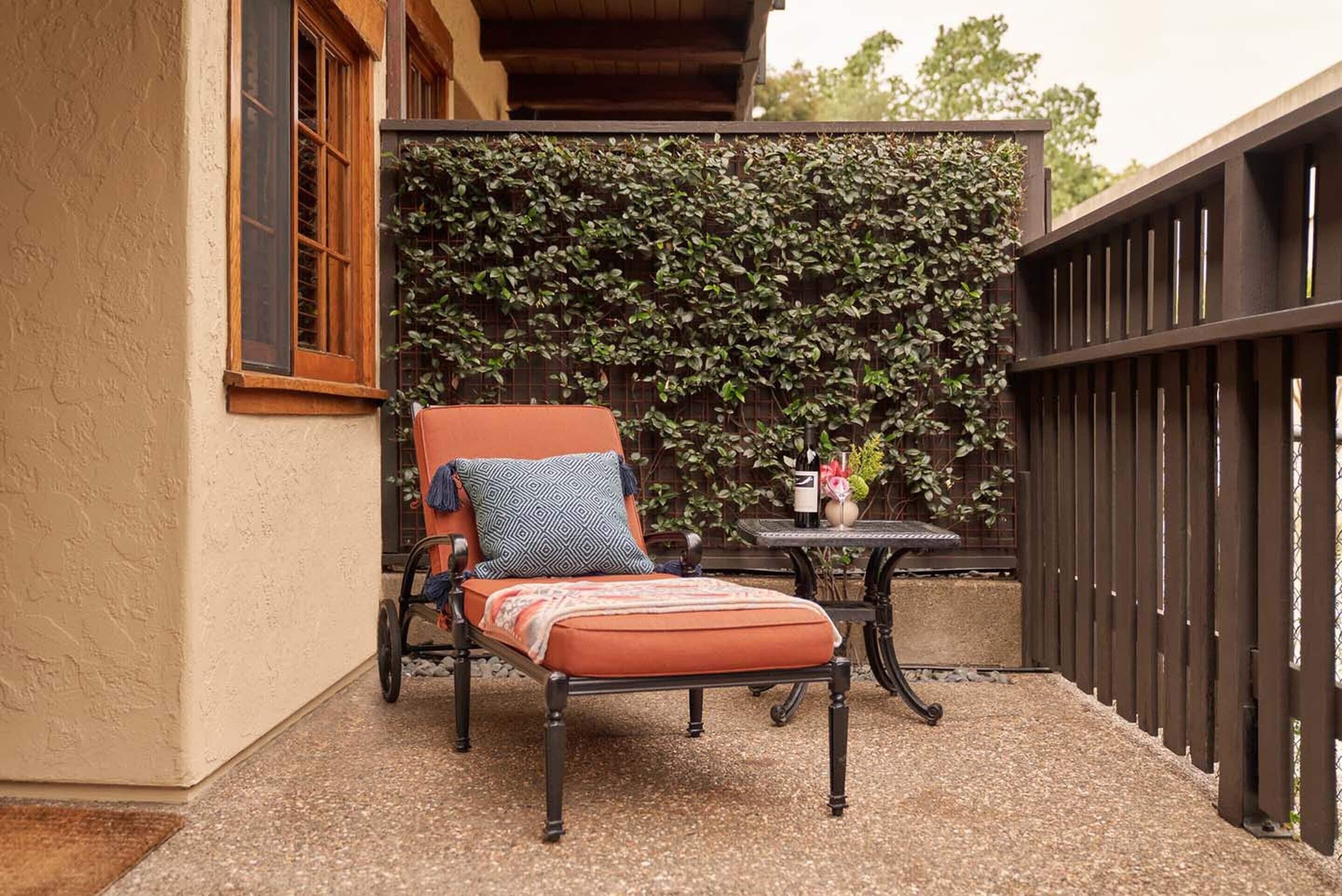 Un espacio al aire libre con acceso para sillas de ruedas, que incluye una silla acolchada y una mesita pequeña, una pared decorativa con plantas trepadoras y una valla de madera.