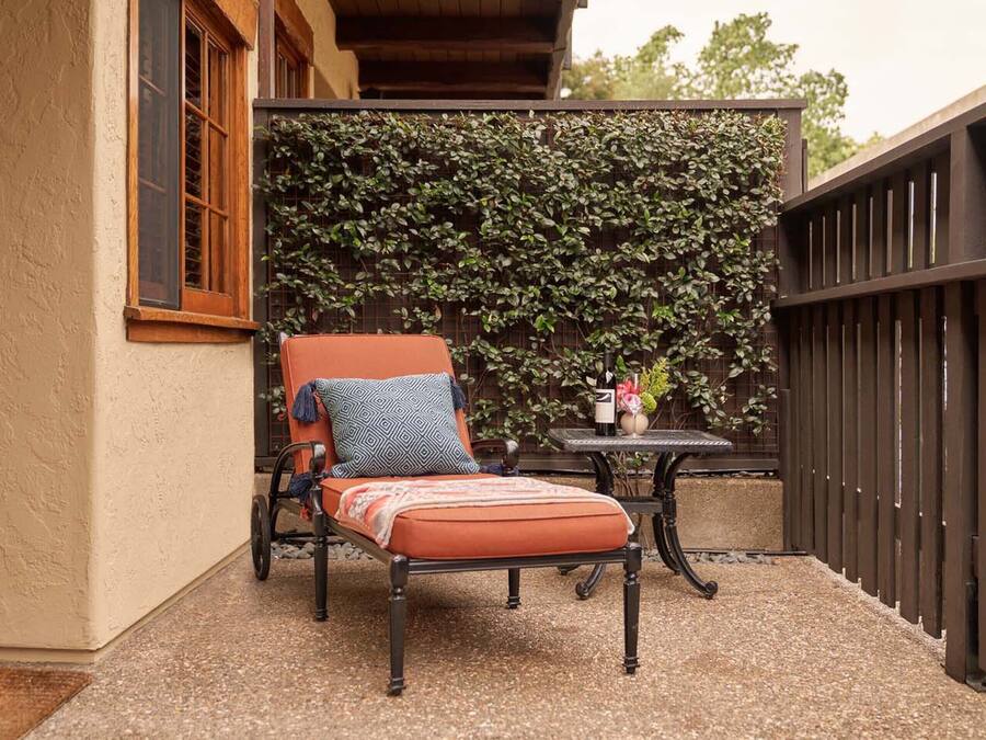 Un espacio al aire libre con acceso para sillas de ruedas, que incluye una silla acolchada y una mesita pequeña, una pared decorativa con plantas trepadoras y una valla de madera.