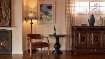 Una stanza accogliente con camino, una sedia di legno, un tavolo con bottiglia e bicchiere, e un mobile contenente oggetti decorativi.
