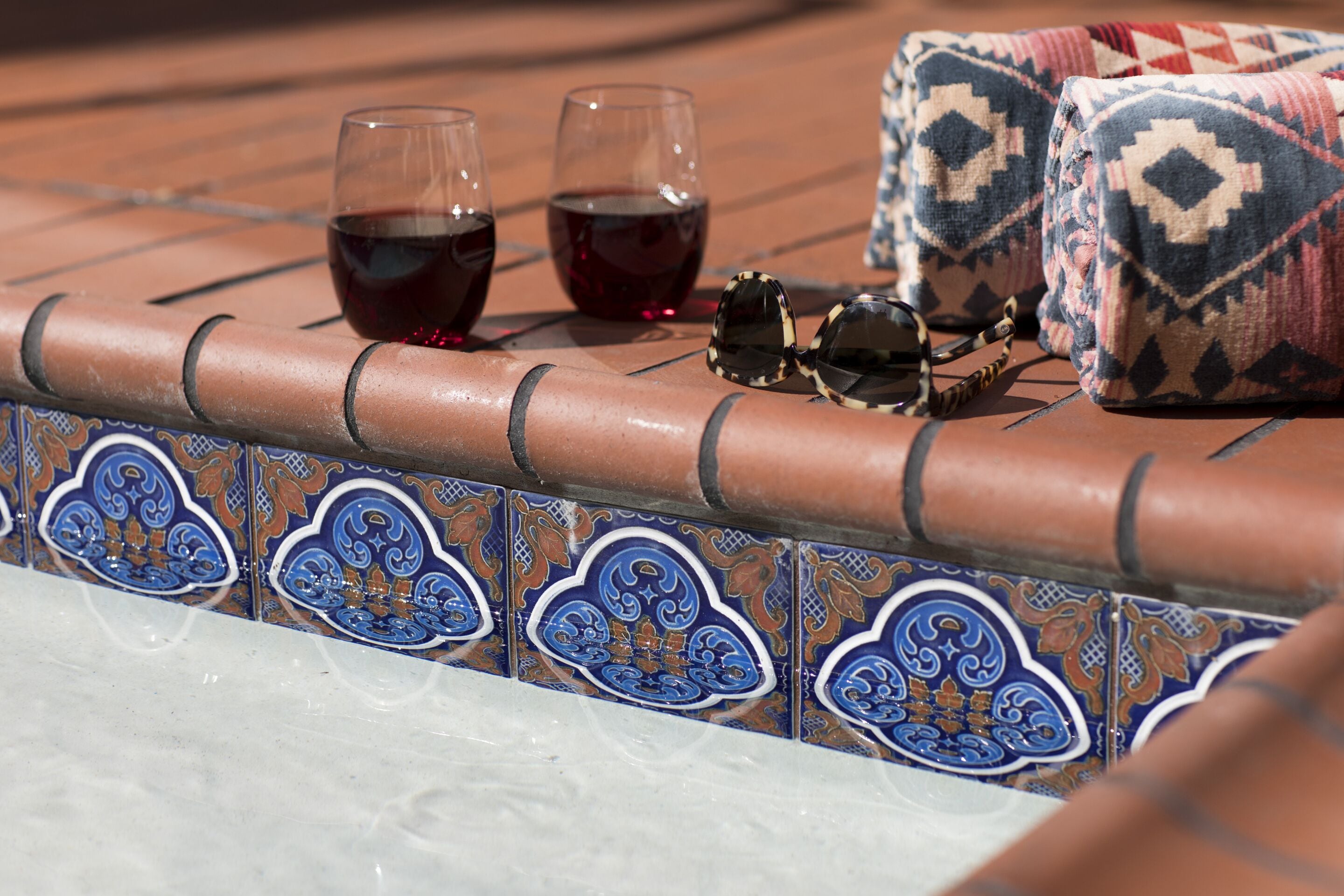 A tiled patio with two glasses of red wine, a pair of sunglasses, and a patterned cushion.