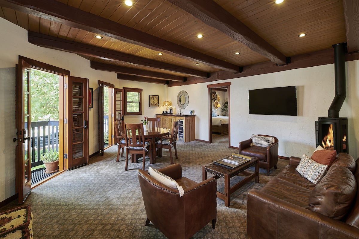 A spacious living room with leather sofas, a coffee table, and a fireplace. There is a dining area with wooden chairs and a table. The room has wooden beams on the ceiling and large windows.