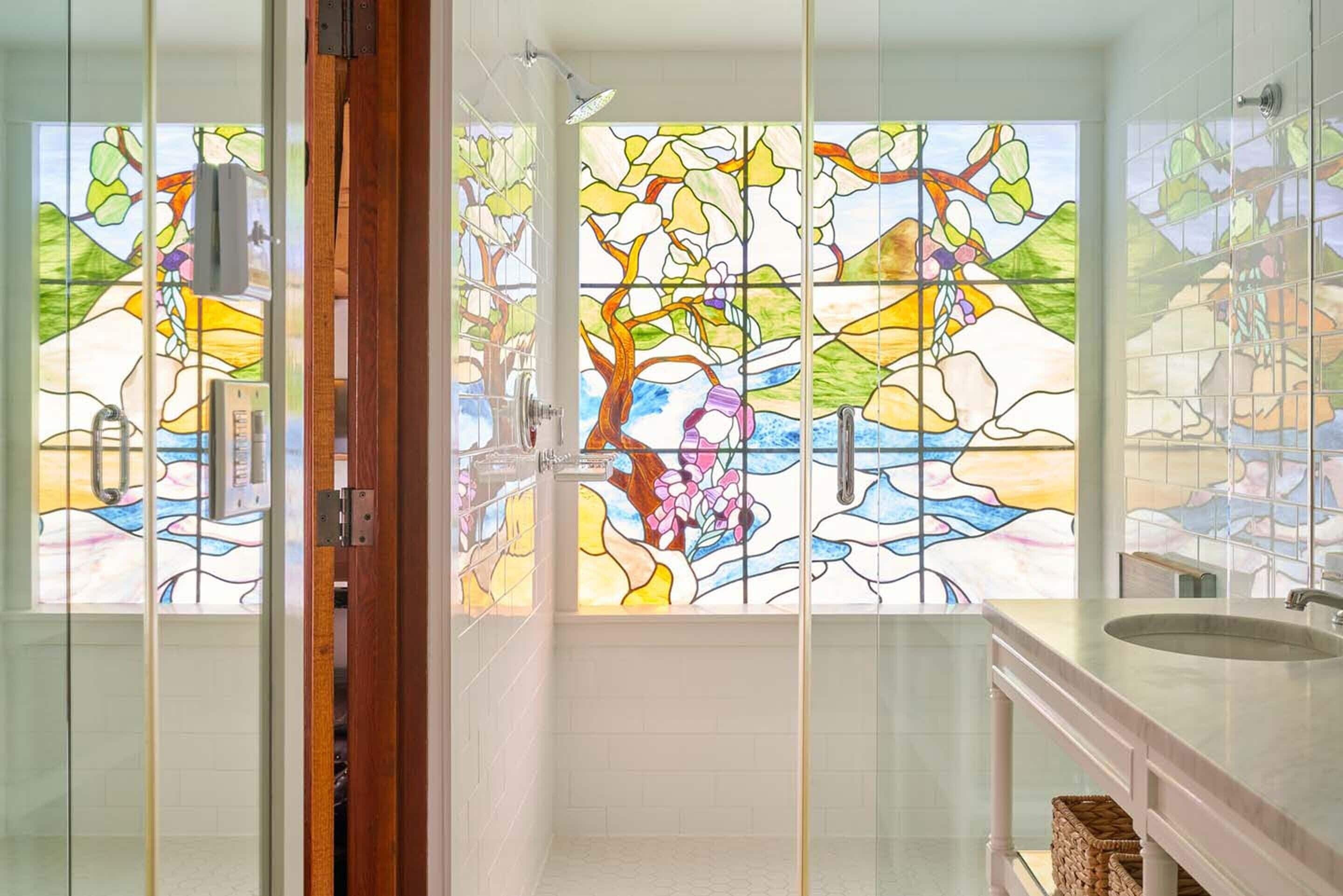 A bathroom with a glass shower enclosure, a sink, and a stained glass window depicting a floral scene.