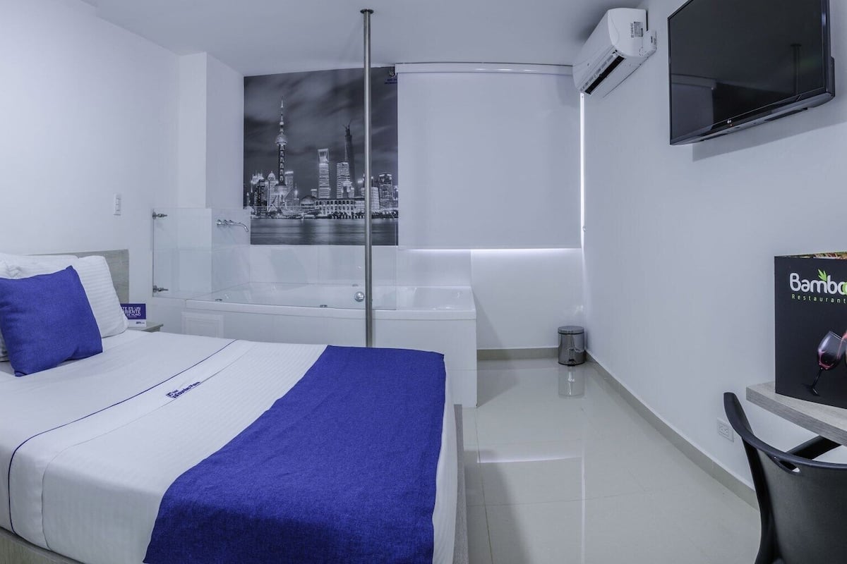 Ein Hotelzimmer mit Bett, Nachttisch, Telefon, Fernbedienung, Badewanne, Dusche, Toilette, Mülleimer, einem an der Wand befestigten Fernseher und Blick auf die Stadt.
