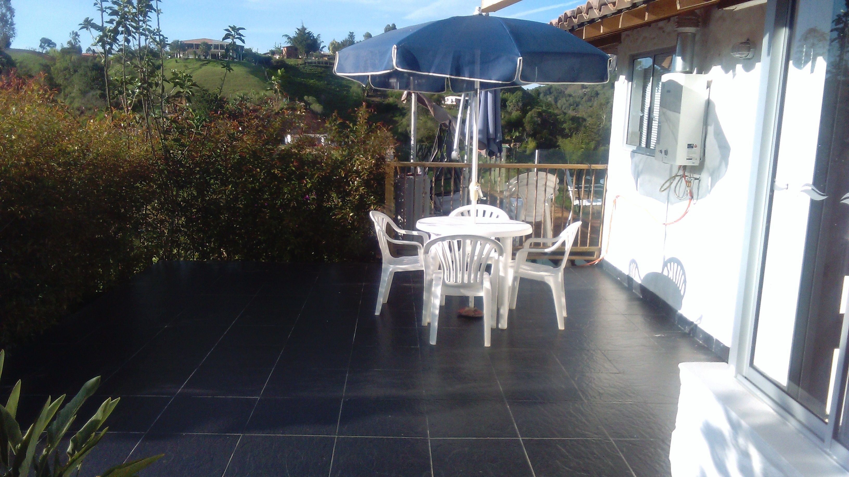 A patio with a table and chairs under a blue umbrella, surrounded by greenery and a view of hills.
