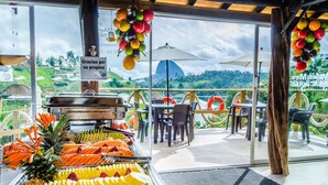A buffet with tropical fruits and vegetables, outdoor seating with umbrellas, and a scenic view of hills and a dome-shaped building. - Hotel Santa Maria de las Aguas Peñol (El Peñol)