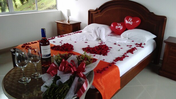 Cama bem arrumada com pétalas de rosa e um balão em formato de coração, uma garrafa de vinho e um buquê sobre uma mesa de cabeceira.