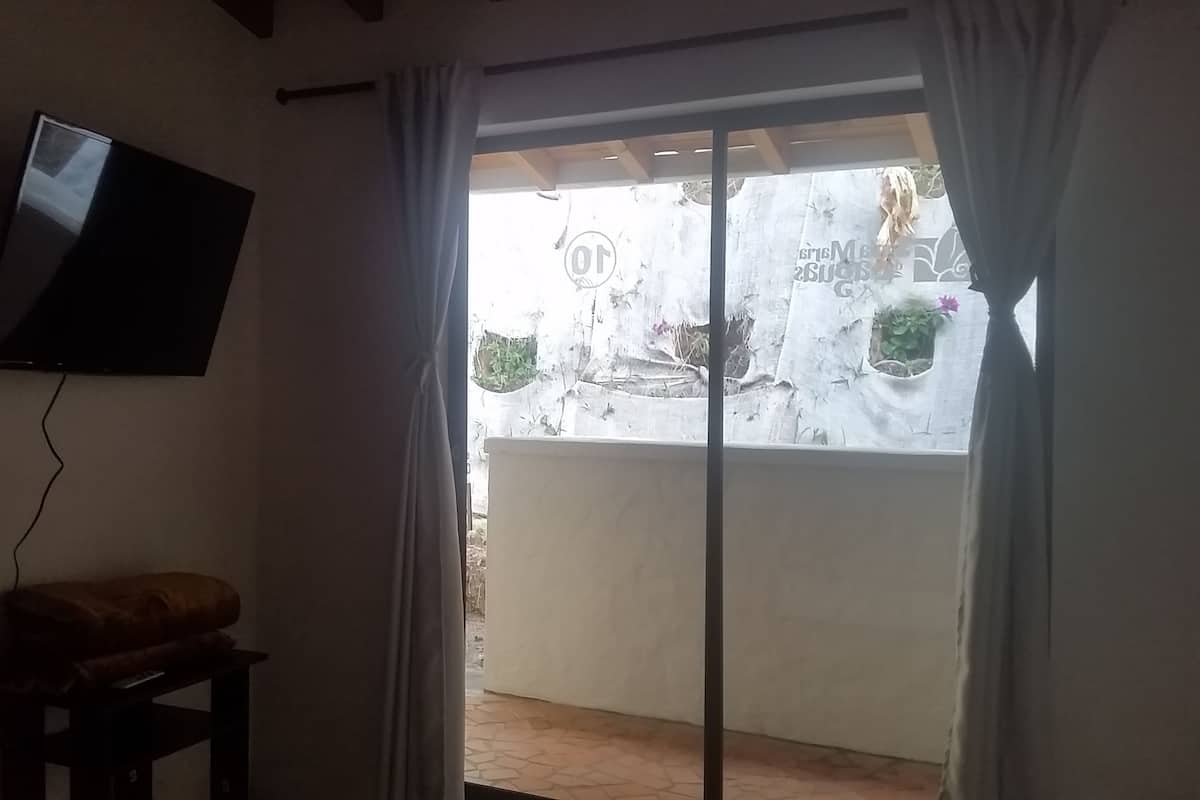 A room with a TV mounted on the wall, a bench, and a sliding glass door leading to an outdoor area with a view of the sky and some greenery.