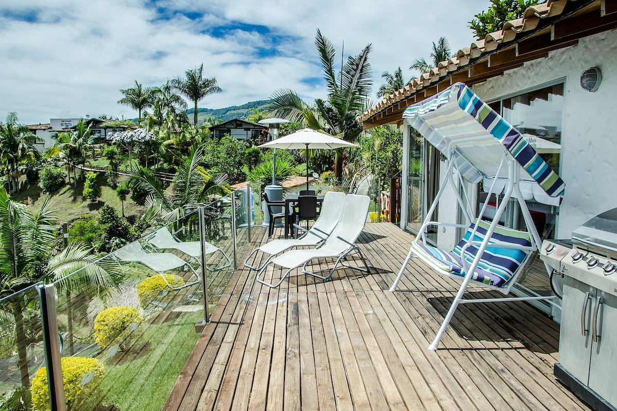 A wooden deck with lounge chairs and a table, surrounded by lush greenery and a view of hills.