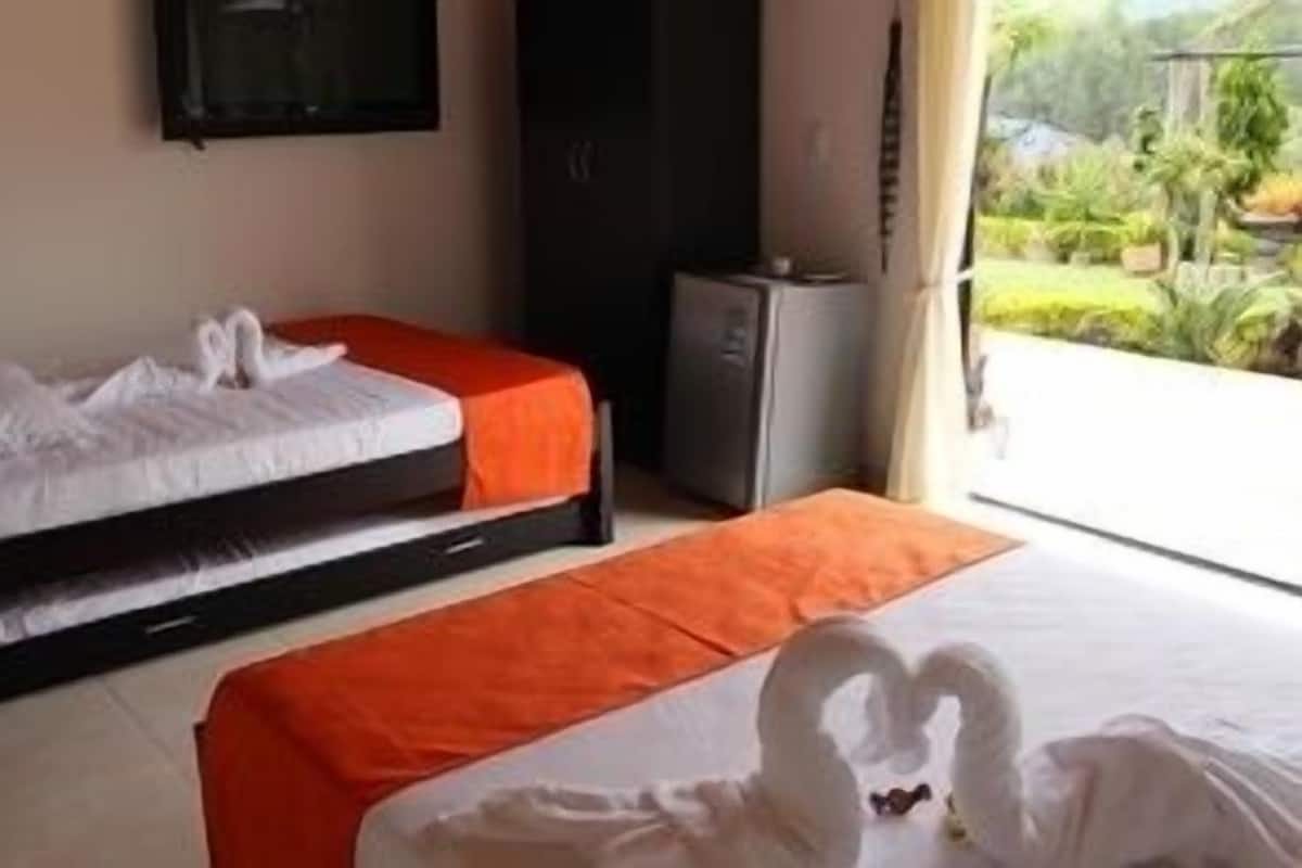 A hotel room with two beds, one with a white bedspread and the other with an orange bedspread. There is a small desk with a computer and a window with a view of greenery outside.