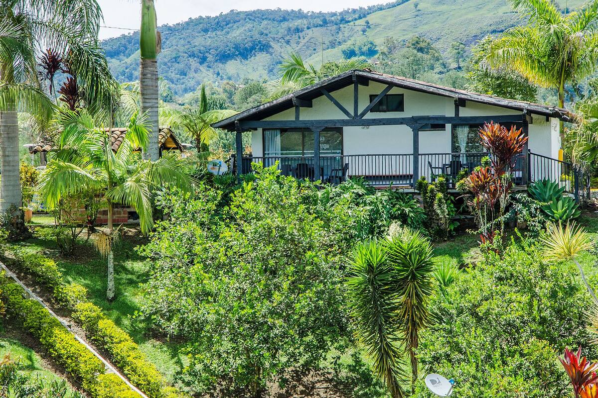 A two-story house with a balcony, surrounded by lush greenery and hills in the background.