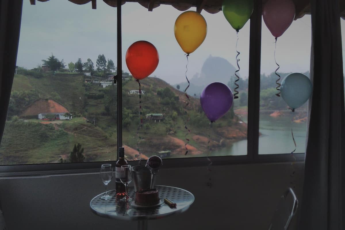 A table with a bottle, wine glasses, and a small cake, with balloons hanging from the window overlooking a rural landscape.