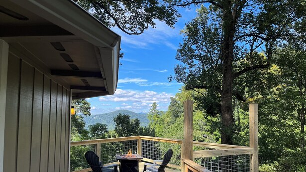 Una terraza de madera con una mesa y sillas, con vistas a una zona boscosa y montañas distantes.
