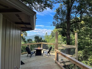 A wooden deck with a table and chairs, overlooking a forested area and distant mountains.