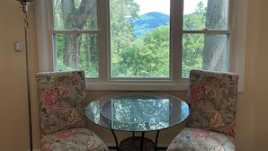 Una habitación con una mesa redonda de vidrio y dos sillones tapizados con motivos florales, ubicados frente a un ventanal con vistas a una ladera boscosa.