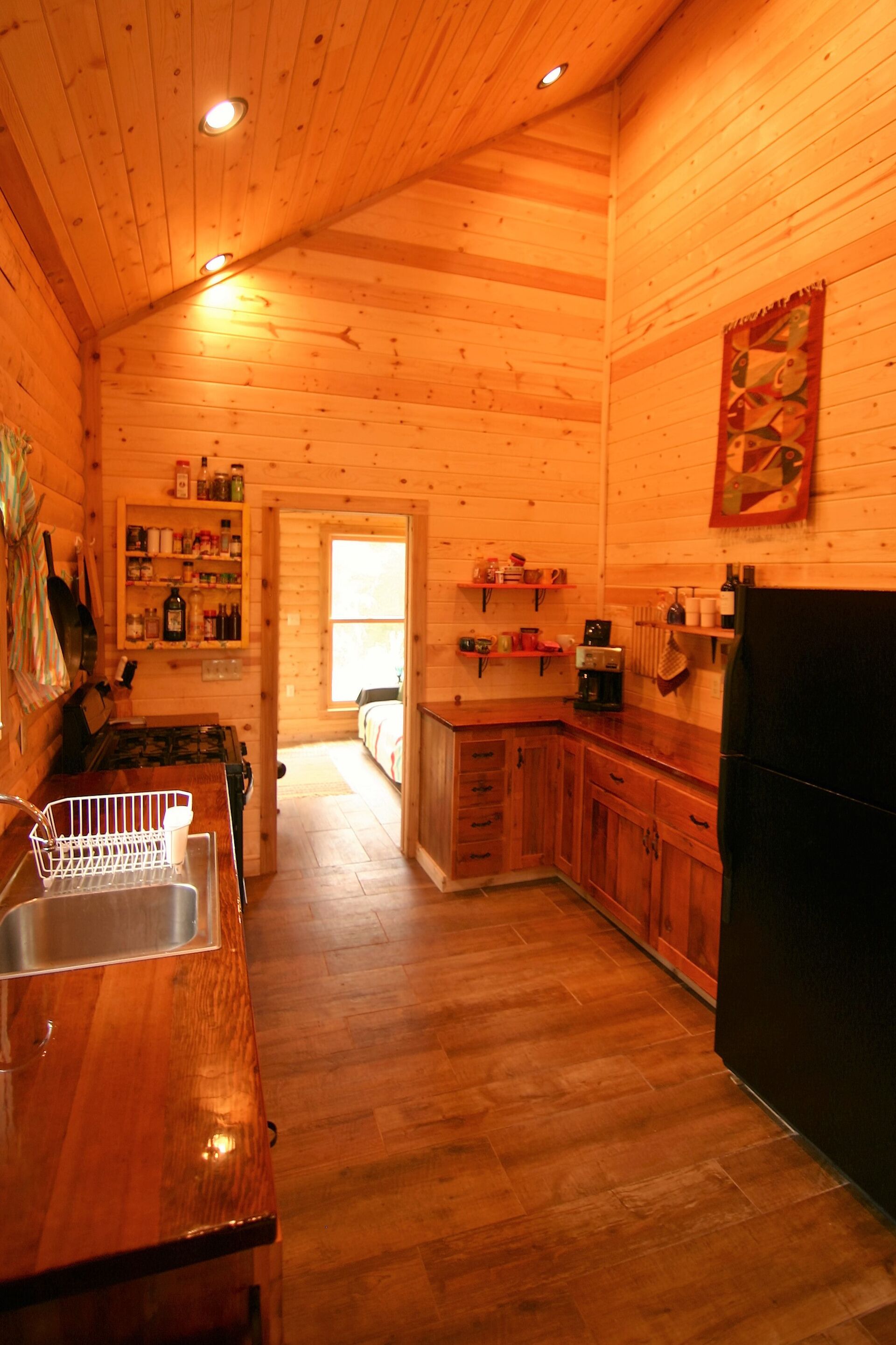 A wooden cabin interior with a kitchen area featuring wooden cabinets, a black refrigerator, and a wooden countertop with a sink.