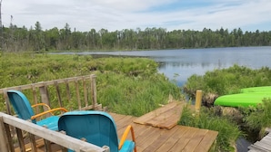 A wooden deck with two blue chairs and a green kayak, overlooking a lake with a forested shoreline.