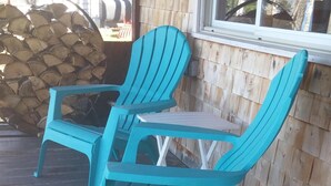 Zwei türkisblaue Adirondack-Stühle und ein kleiner Tisch auf einer Holzterrasse mit Blick auf Bäume und ein Gebäude im Hintergrund.