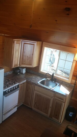 A compact kitchen with wooden cabinets, a built-in stove, a sink, and a window providing natural light. - Brassua Lake Camps in Rockwood, Maine (Antler) (Rockwood)