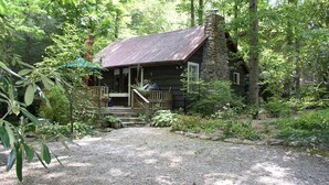 A rustic cabin with a stone chimney, wooden deck, and a gravel driveway surrounded by dense forest.