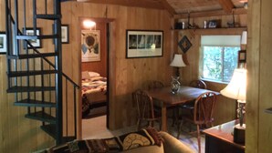 A cozy cabin interior with a wooden staircase, a dining area with a table and chairs, and a bed in the background.