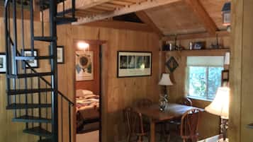 A cozy cabin interior with a wooden staircase, a dining area with a table and chairs, and a bed in the background.