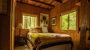 A wooden cabin bedroom with a bed, two windows, a nightstand, and a lamp.