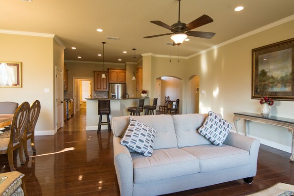 A spacious living room with a grey sofa, wooden chairs, a dining table, and a kitchen in the background.