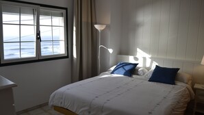 Een slaapkamer met een bed, nachtkastje, lamp, raam met gordijnen en uitzicht op zee.
