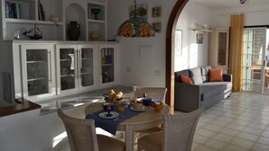 Een eetgedeelte met een ronde tafel, stoelen en een ontbijtopstelling. Er is een witte kast met glazen deuren en een hangende decoratieve lamp.